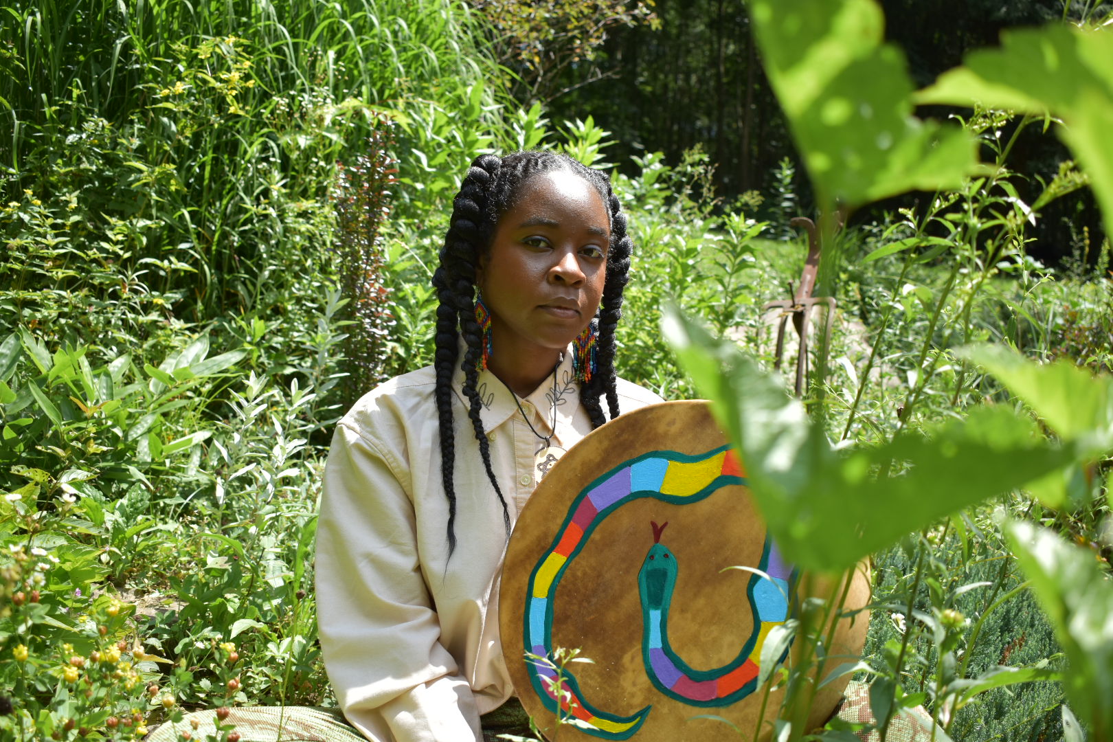 Dark brown skinned nonbinary person sits in a green summer garden. They are wearing a white button up shirt and holding a hand drum with a painted rainbow snake.