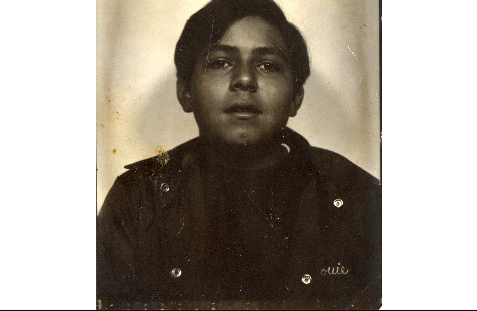 The image depicts a teen boy. The images is black and white. He is wearing a windbreaker with Louie stiched on it. His has a dimple on his chin, which is lightly tilted up. He is looking at the camera. 