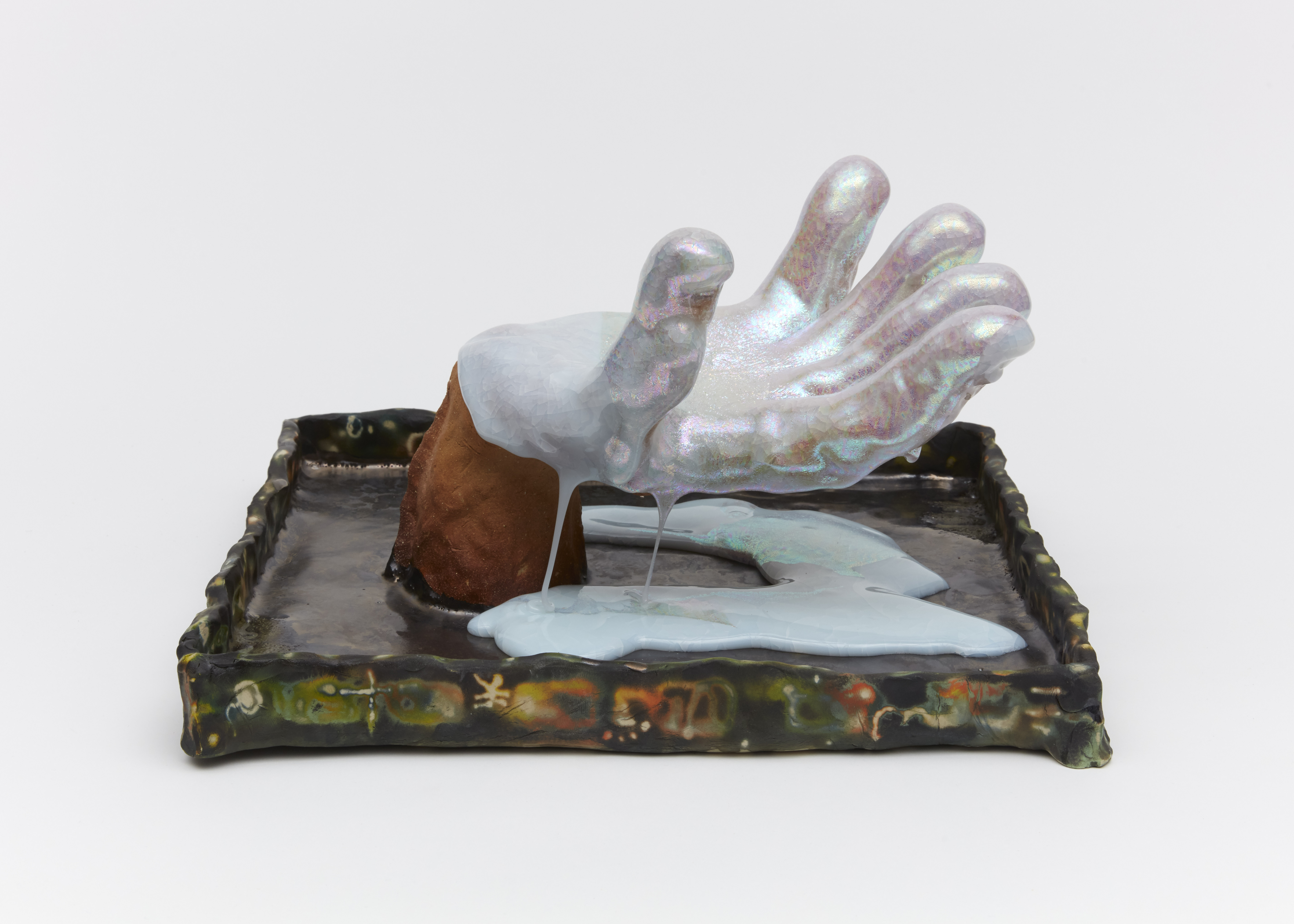 Hand (slipping through) is a sculptural hand made of porcelain inside of a square “tray”. The hand is covered with melted, milky white, iridescent glass, which drips and slips through the fingers. The hand is made to look as if it is reaching through the tray, attempting to grasp something. The tray is filled with metallic glazes and has hieroglyphic type designs on the outside. 
