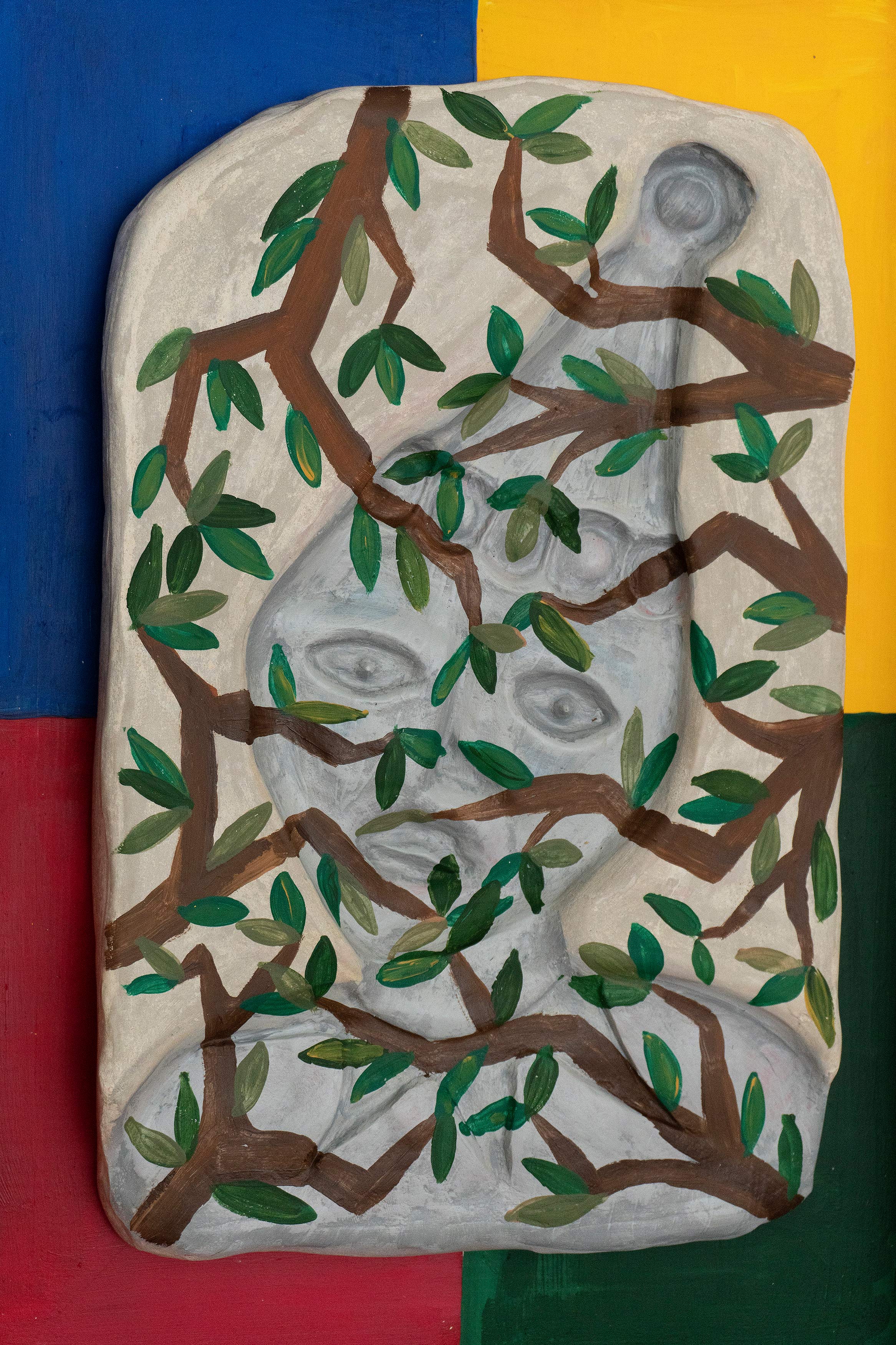A grey hydrocal cast of a clown rests in the middle of a wooden frame. The cast has painted foliage on it, obscuring the image of the clown. The wooden frame is painted in quarters; blue, yellow, green and red. The frame has a pastel diamond harlequin border.
