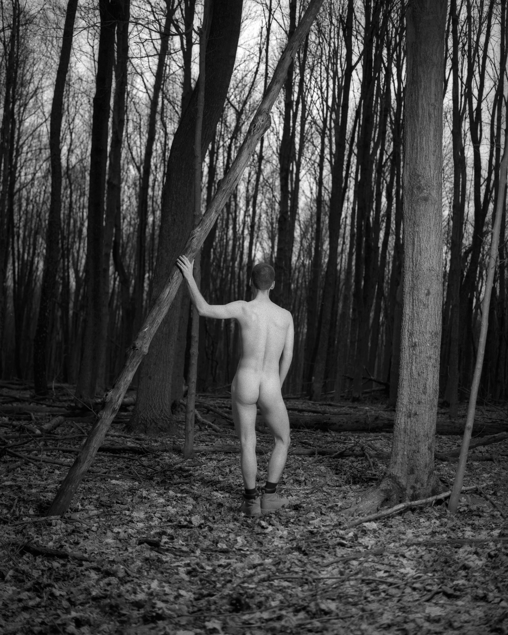 Black and white nude self portrait in the woods behind my house in Ohio. I’m standing contrapposto with my back to the camera while gripping a tree that has fallen with my left hand. 
