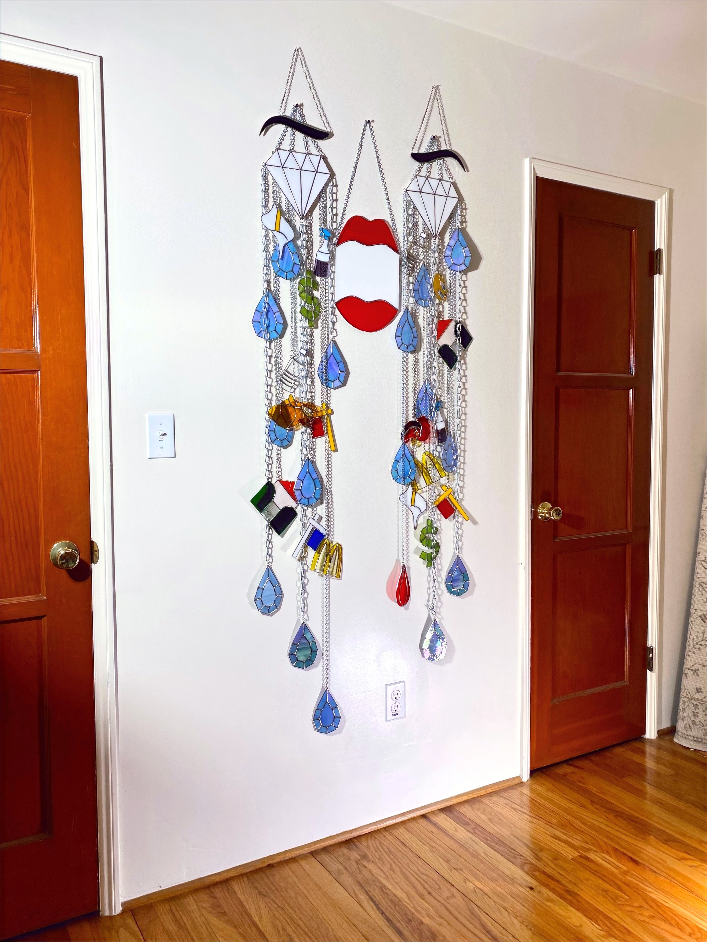 A large stained glass work hangs in an apartment bedroom. The work visualizes dark eyebrows, diamonds for eyes, an agape mouth, and a flow of teardrop gem tears amongst assorted icons, all bound by chains.